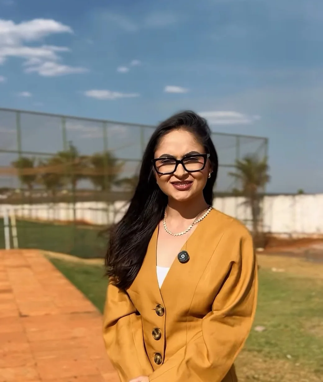 Andressa Miranda sorrindo, de terno mostarda, colar de pérolas e óculos, ao ar livre sob o céu azul do cerrado.