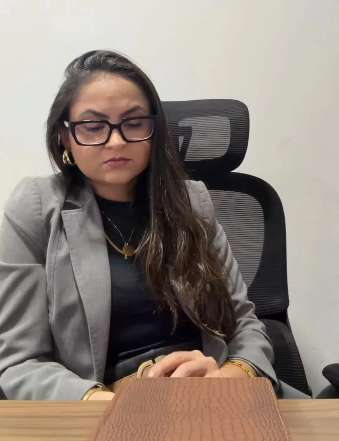 Andressa Miranda à mesa do escritório, de blazer cinza e óculos, lendo um documento sobre uma pasta de couro.
